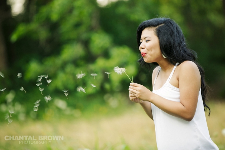 Dallas senior photographer a portrait of Lien blowing dandelions at Richardson Breckinridge Park surrounded by vivid green grass by Chantal Brown Photography.