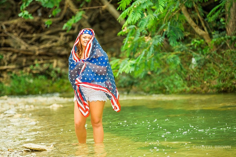 Laughing and having fun with Dallas senior portraits photographer of a Mesquite high school student wrapped around American flag scarf and standing in the water creek river.